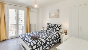 A bedroom with a bed, bedside tables, a desk, a hanging light fixture, a window with curtains, and a wall-mounted picture.