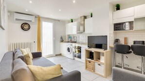 A modern living room with a grey sofa, a flat-screen TV, and a kitchen area with white cabinets and a microwave.