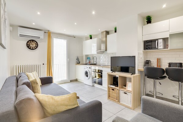 Una sala de estar moderna con un sofá gris, un televisor de pantalla plana y una zona de cocina con armarios blancos y un microondas.