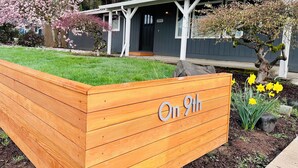 Un bac à fleurs en bois orné de l’inscription « On 9th », un jardin planté de jonquilles et un bâtiment doté d’un porche.