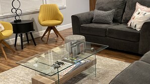 Un salon avec une table basse en verre, un canapé gris, un fauteuil jaune et une table d’appoint noire.