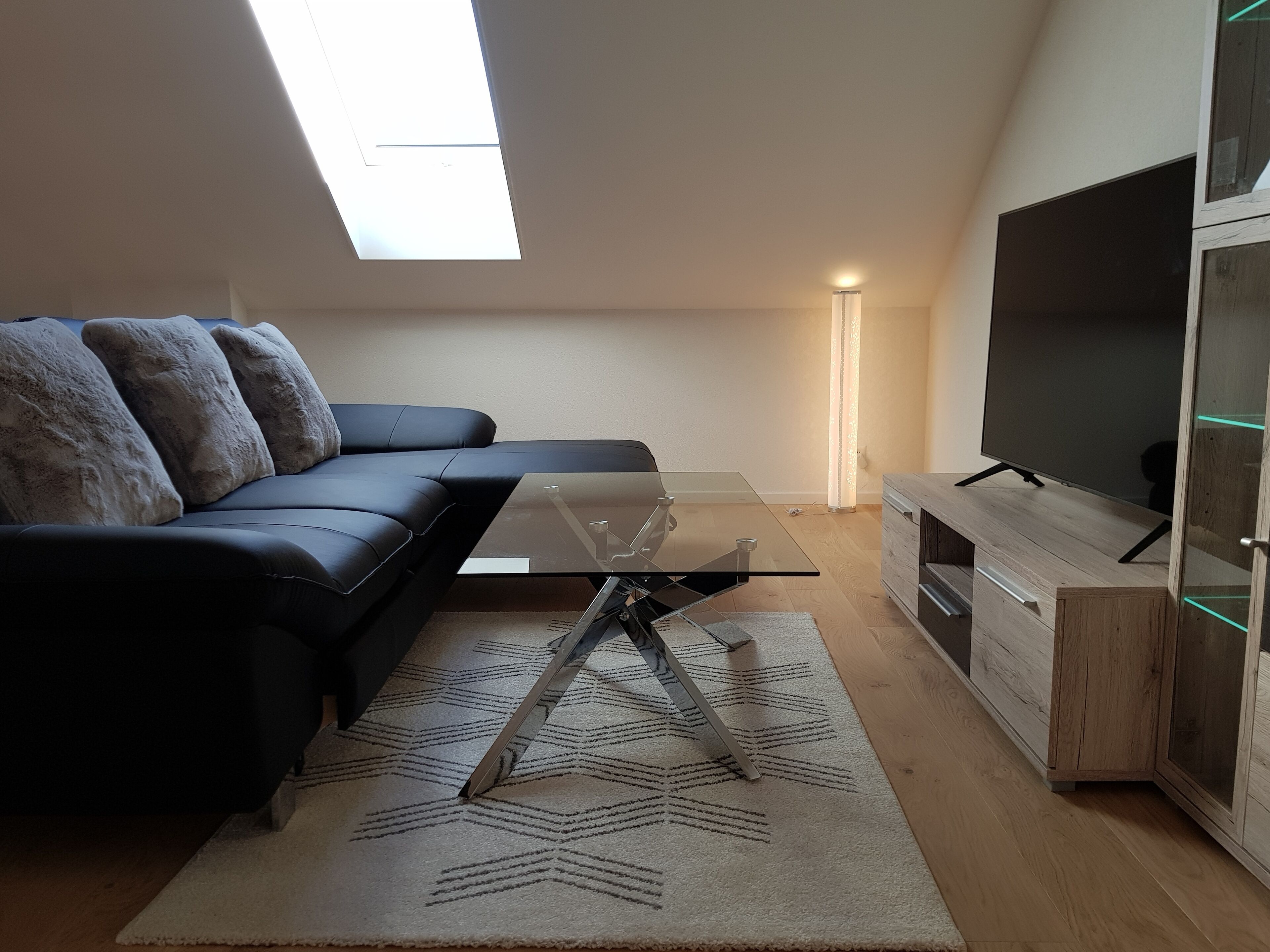 A modern living room with a glass-top coffee table, a dark leather sofa, a patterned rug, a wooden cabinet with a TV, and a skylight.