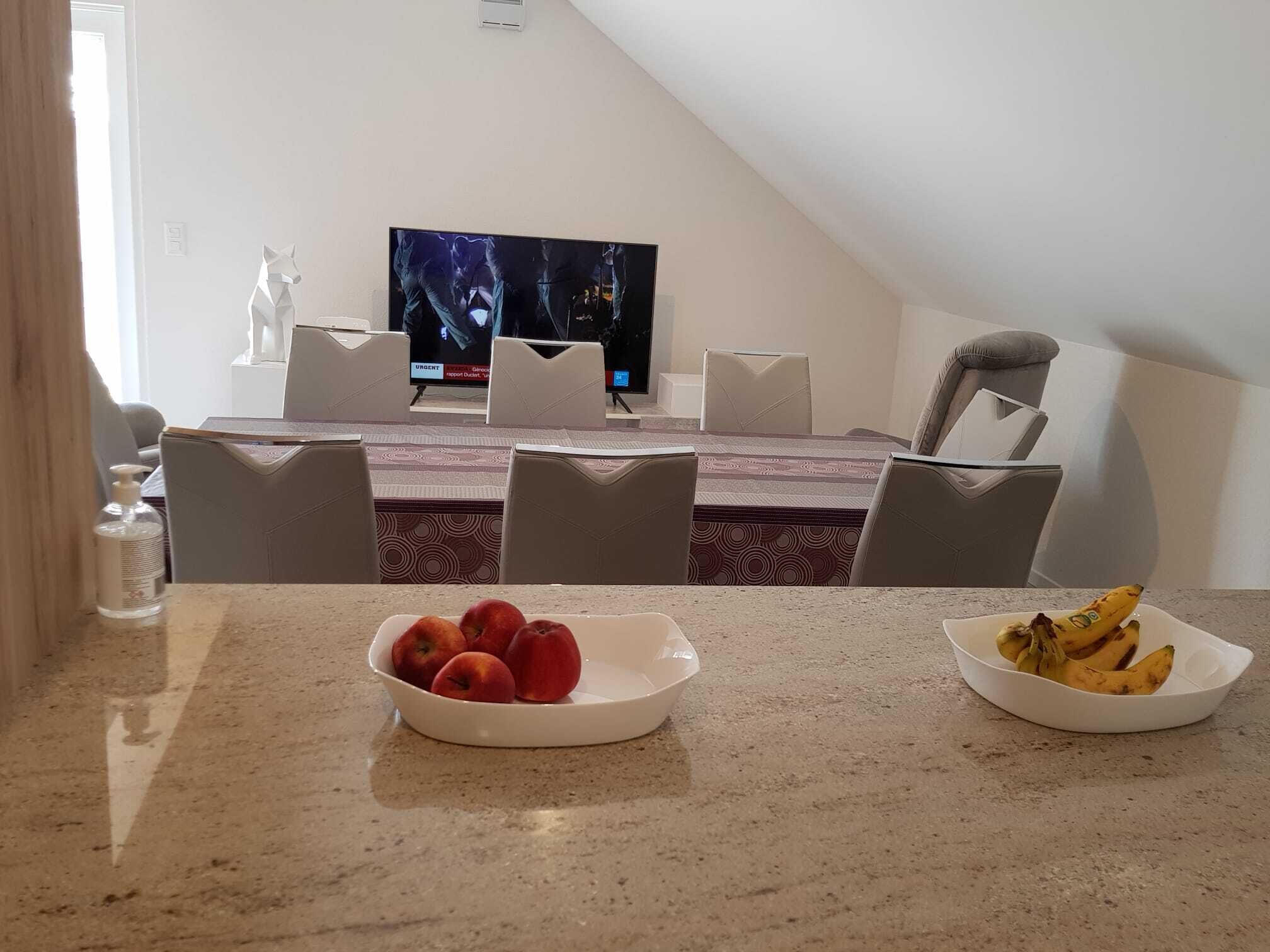 A dining area with a table set for four, a television in the background, and a bowl of fruit on the table.