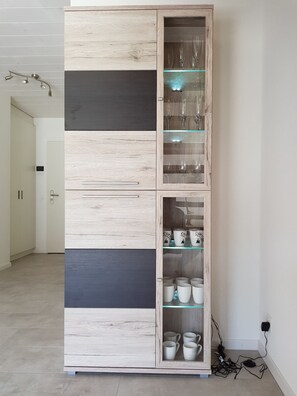 A tall, multi-paneled cabinet with a mix of wood and dark panels, containing glass shelves with wine glasses and coffee mugs.