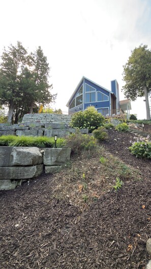 A modern house with a stone retaining wall, a well-maintained garden with mulch and plants, and a clear sky. - 5BR Waterfront Retreat – Experience Door County’s Best Season, Fall! (Sturgeon Bay)