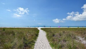 Un sendero en la playa que lleva al océano, con tiendas de campaña y gente a lo lejos.