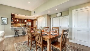 Un comedor con una mesa de madera dispuesta para seis personas, rodeada de sillas de madera. Se observa una cocina con armarios de madera y un refrigerador al fondo.
