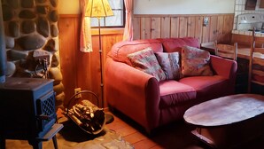 Una sala de estar acogedora con pared de piedra, estufa de madera, sofá rojo con cojines estampados y una pequeña mesa redonda.