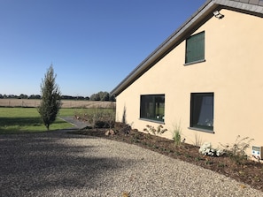 Ein Haus mit Schotterweg, einem kleinen Baum und einem Feld im Hintergrund.