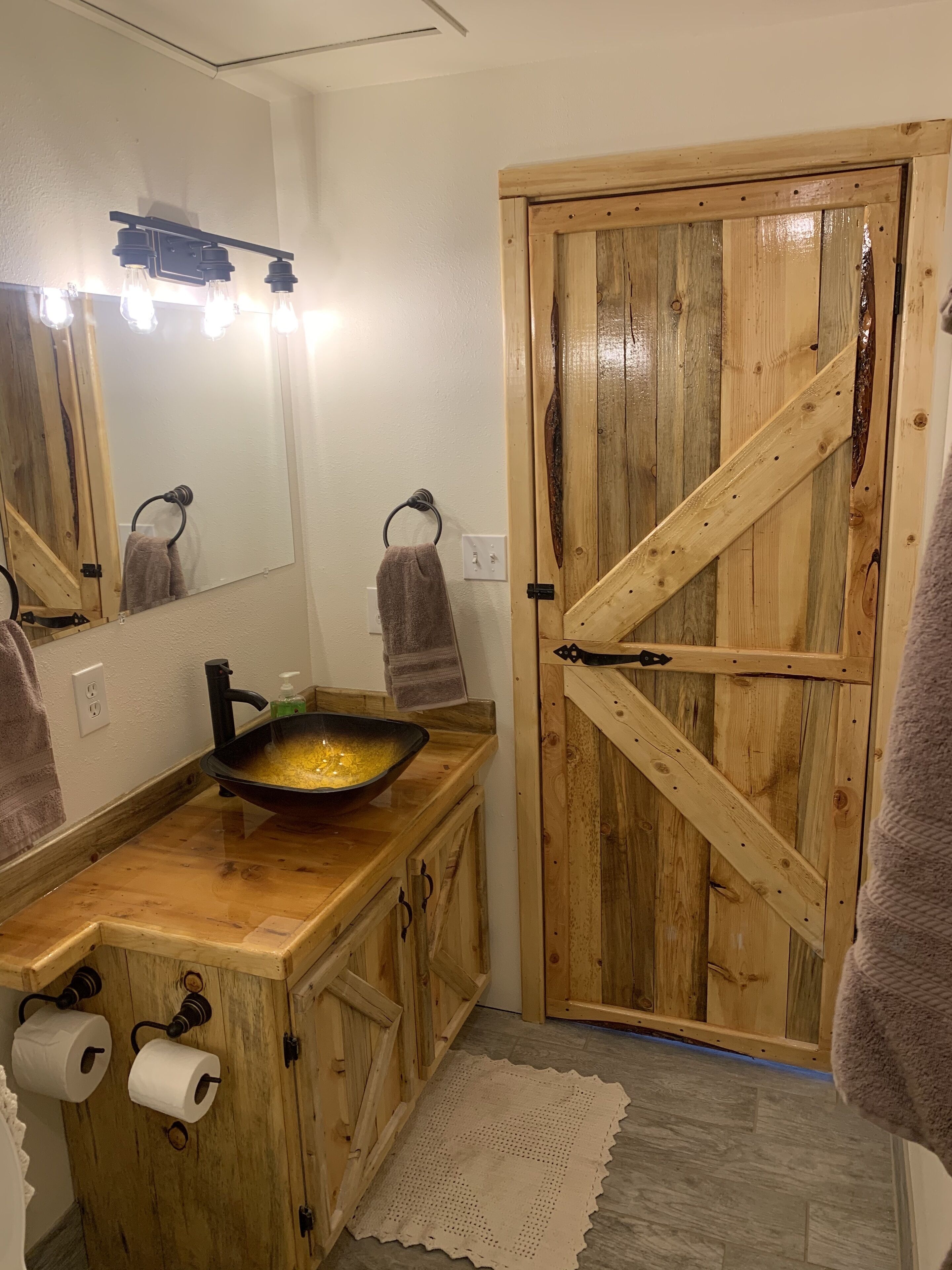 Un baño con tocador de madera, un lavamanos con un recipiente negro, dos toallas, un espejo con luces y una puerta de madera.