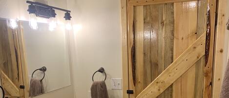 Un baño con tocador de madera, un lavamanos con un recipiente negro, dos toallas, un espejo con luces y una puerta de madera.
