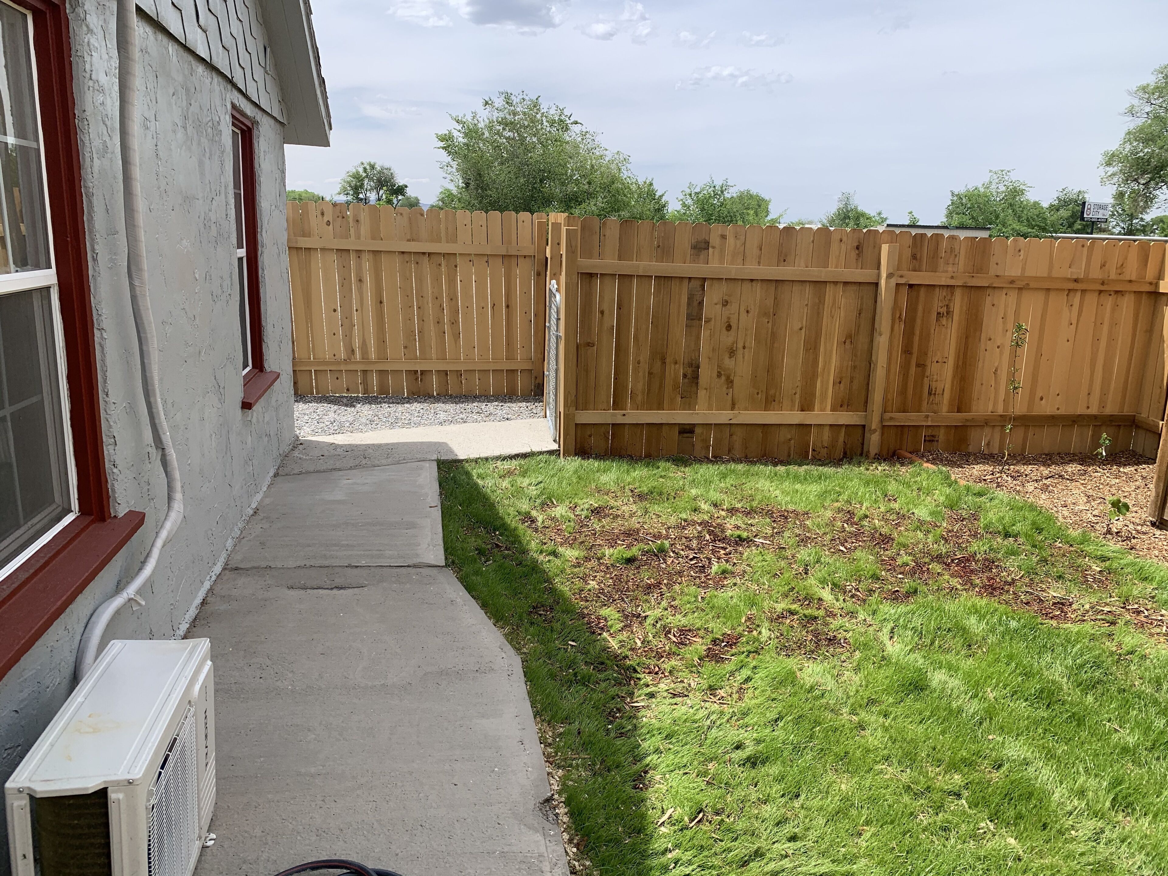 Un patio trasero con cerco de madera, un sendero de concreto y una zona verde con plantas jóvenes.