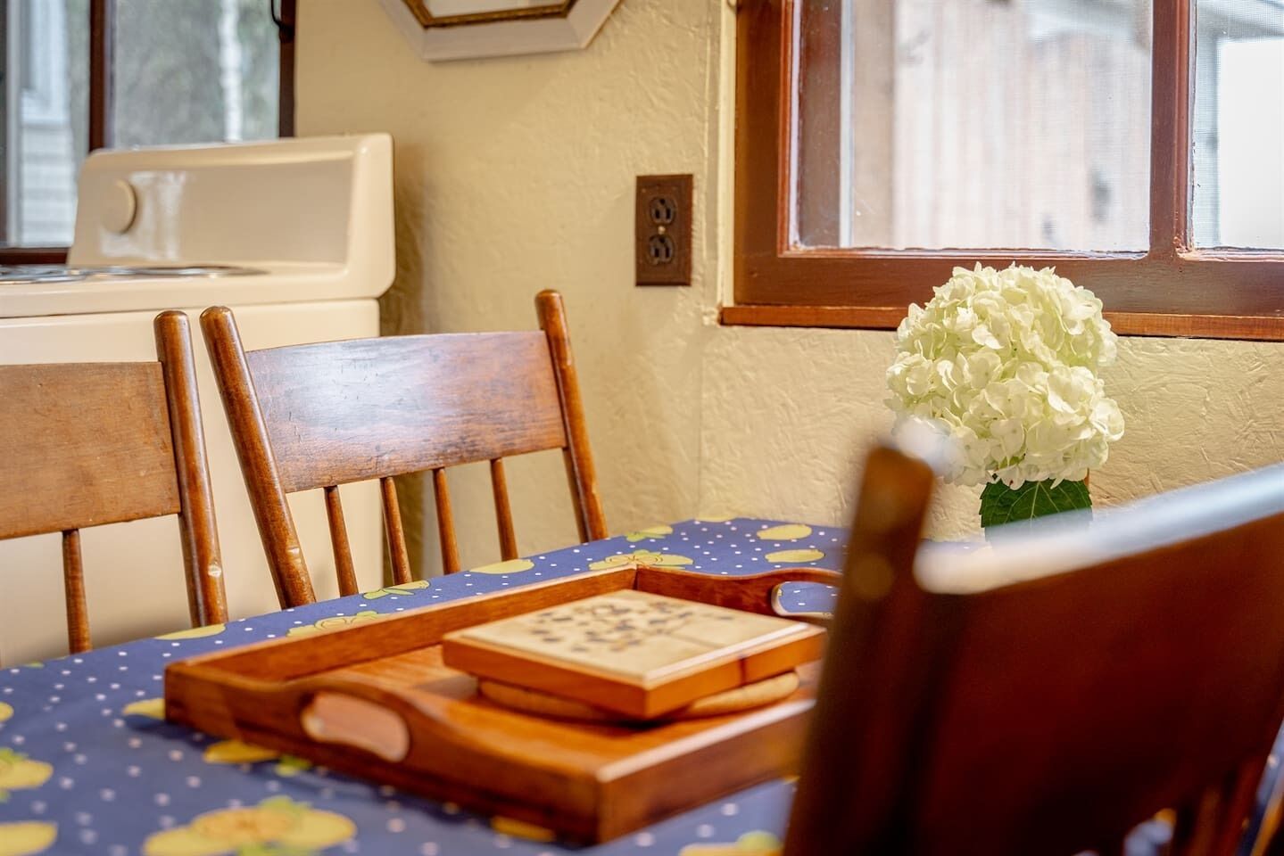 A dining area with a wooden chair, a table with a blue tablecloth, a vase with white flowers, and a wooden tray with a puzzle.
