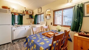 Een compacte keuken met witte kasten, een magnetron en een fornuis. Er is een eettafel met stoelen en een blauwe tafelkleed met citroenmotieven. Een klein houten kastje is rechts zichtbaar.