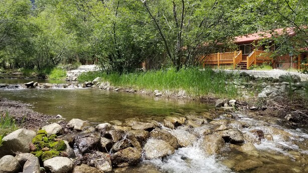 Eine rustikale Holzhütte mit einer Holzdiele und einem Kiesweg, der zu einer Holzbrücke über einen klaren Bach führt.