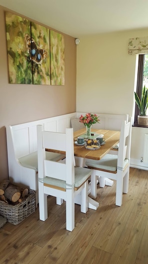 Un coin repas avec une table en bois, des bancs blancs avec coussins, un vase rempli de fleurs et un tableau accroché au mur.