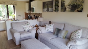 A living room with a grey sofa set, a kitchen area with a stove and oven, and two wall-mounted pictures.