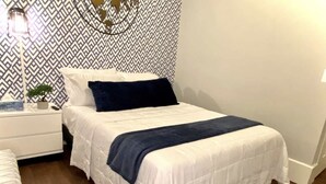 A neatly made bed with a white and blue bedspread, a small bedside table with a lamp, and a patterned wall. - NEW! Cozy Stay in Newly Decorated Apt, in Midtown! (Detroit)
