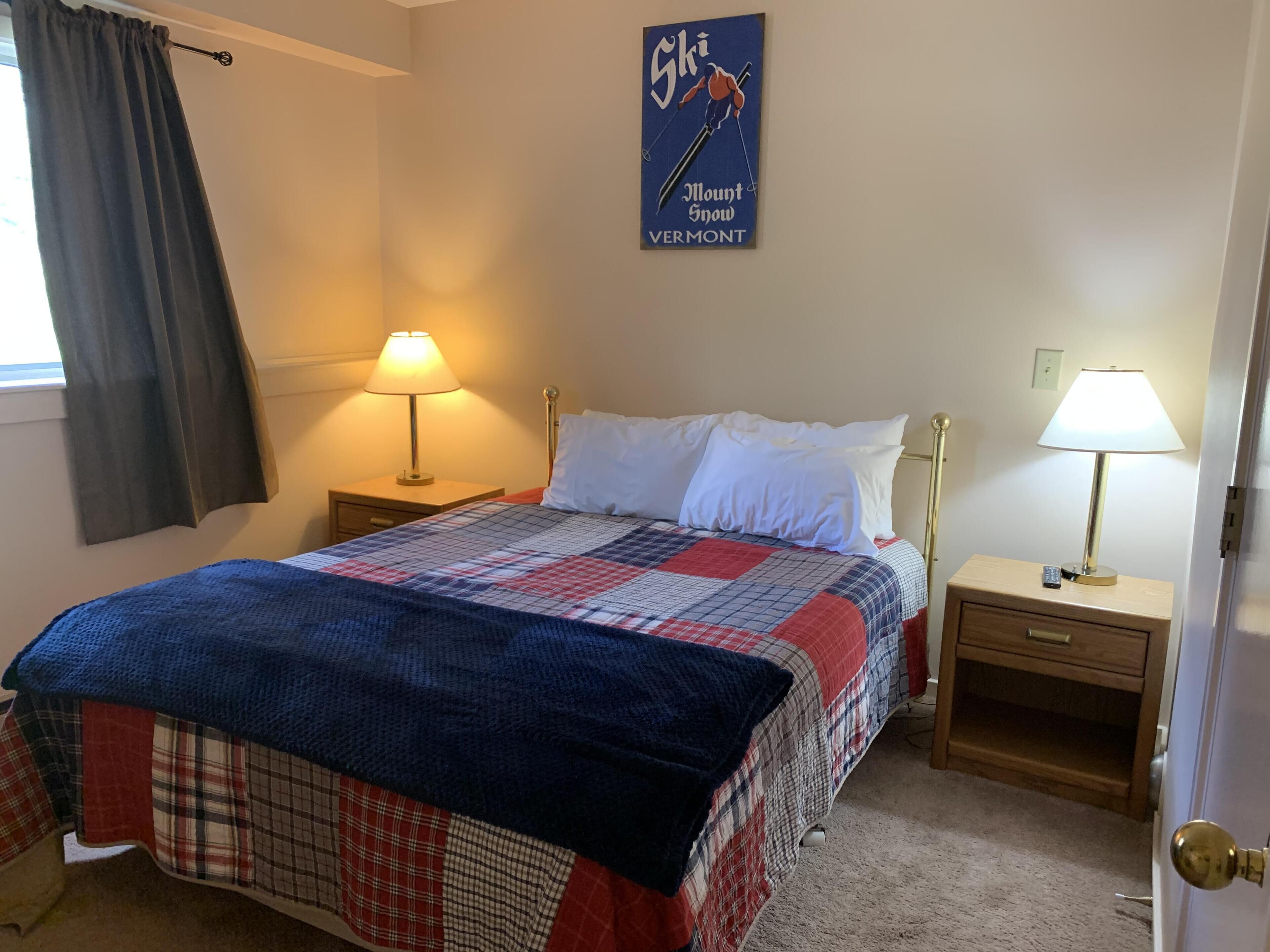 Une chambre à coucher avec un lit, deux tables de chevet avec des lampes, une fenêtre avec des rideaux et une affiche accrochée au mur.