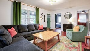 A living room with a dark grey sectional sofa, a wooden coffee table, a green armchair, and a patterned rug. - Deacon Stays: The Brookwood - Walk to WFU (Winston-Salem)
