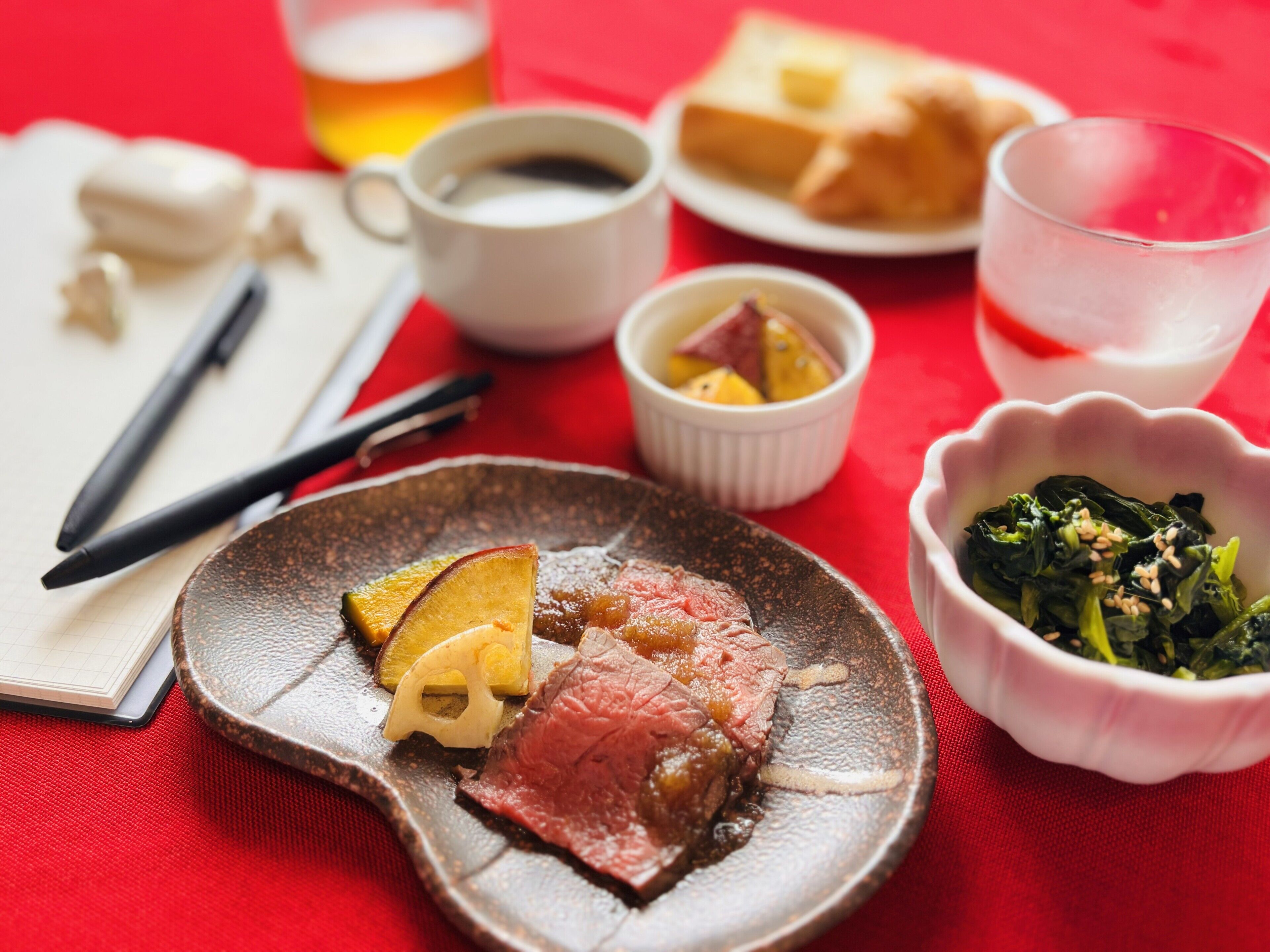 A meal with various dishes including a plate of sliced meat, a bowl of greens, and a cup of tea on a red tablecloth.