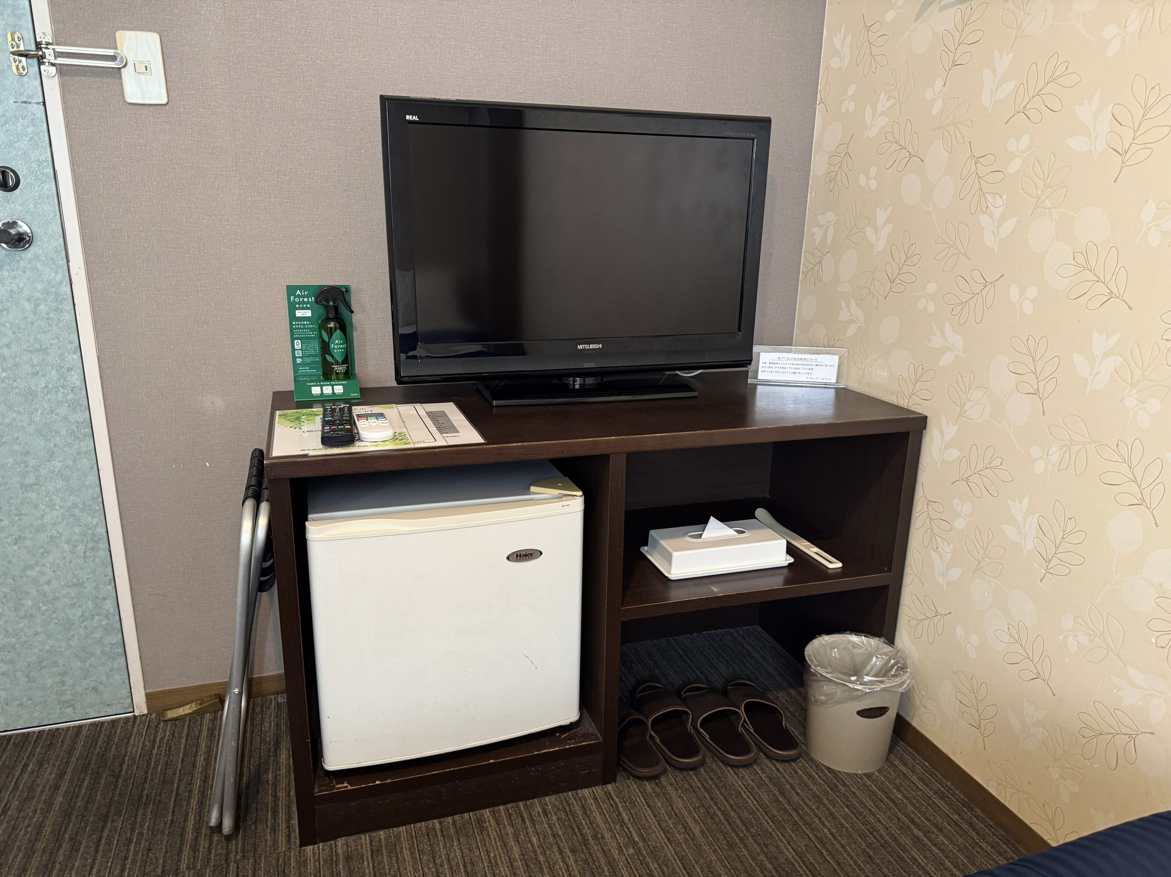A hotel room with a television, a mini-fridge, a trash bin, slippers, and a remote control on a wooden stand.