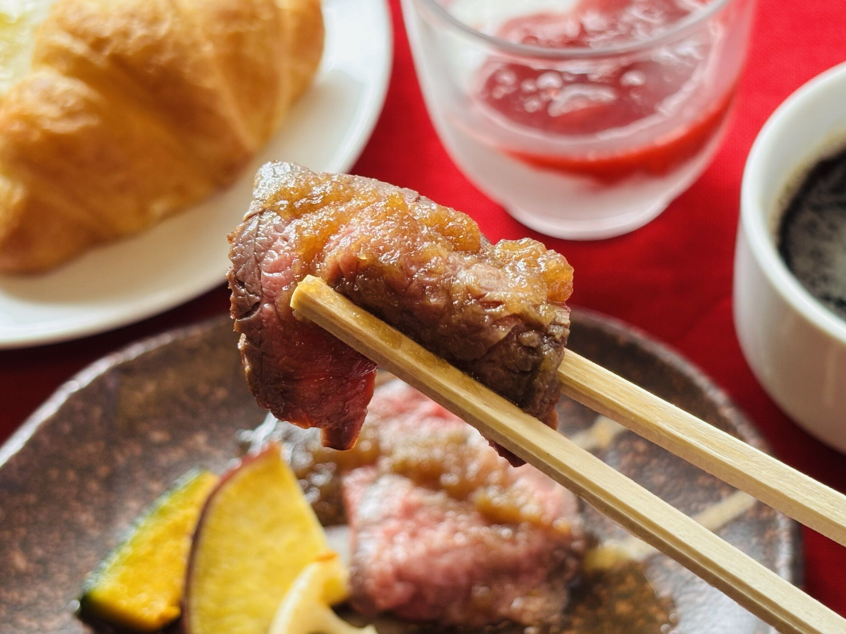 A close-up of a food item being held by chopsticks, with a plate and a glass of red beverage in the background.
