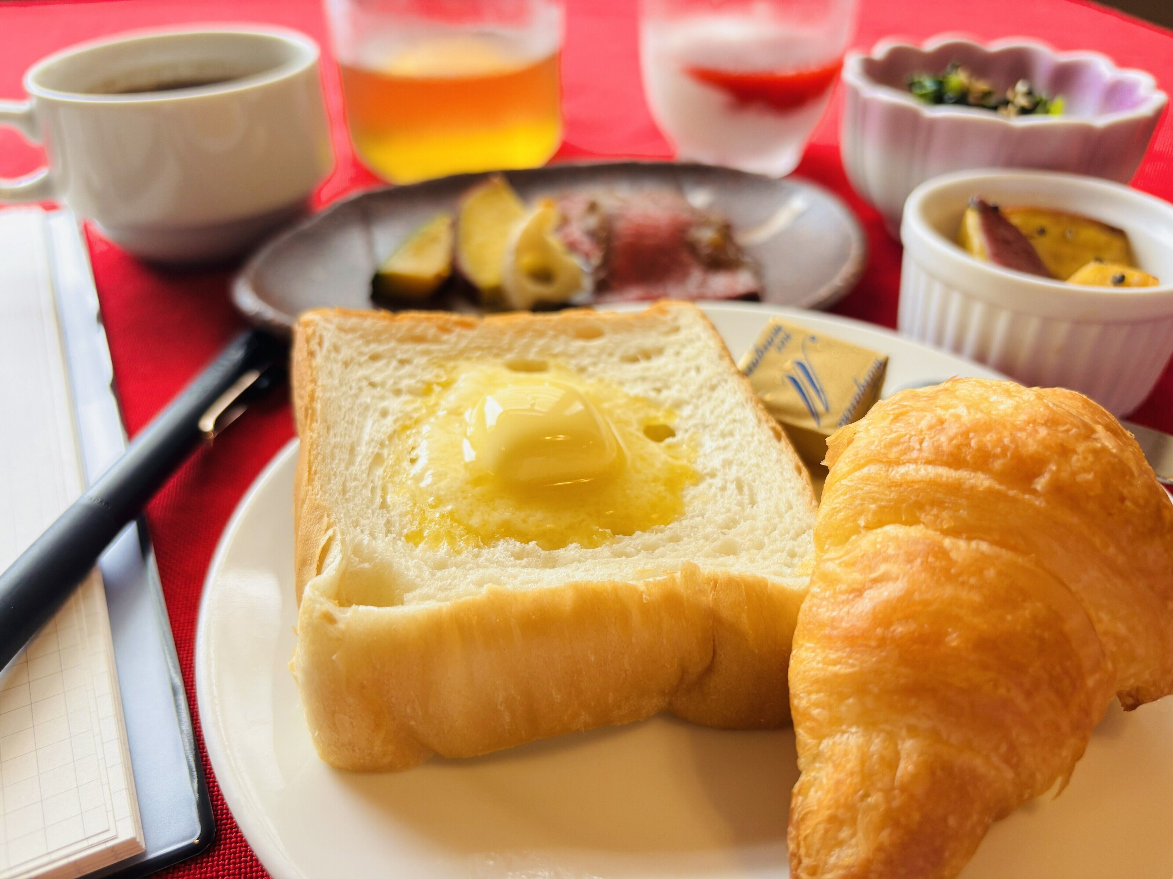 A breakfast setting with toast, butter, a croissant, and small dishes of food on a red table.