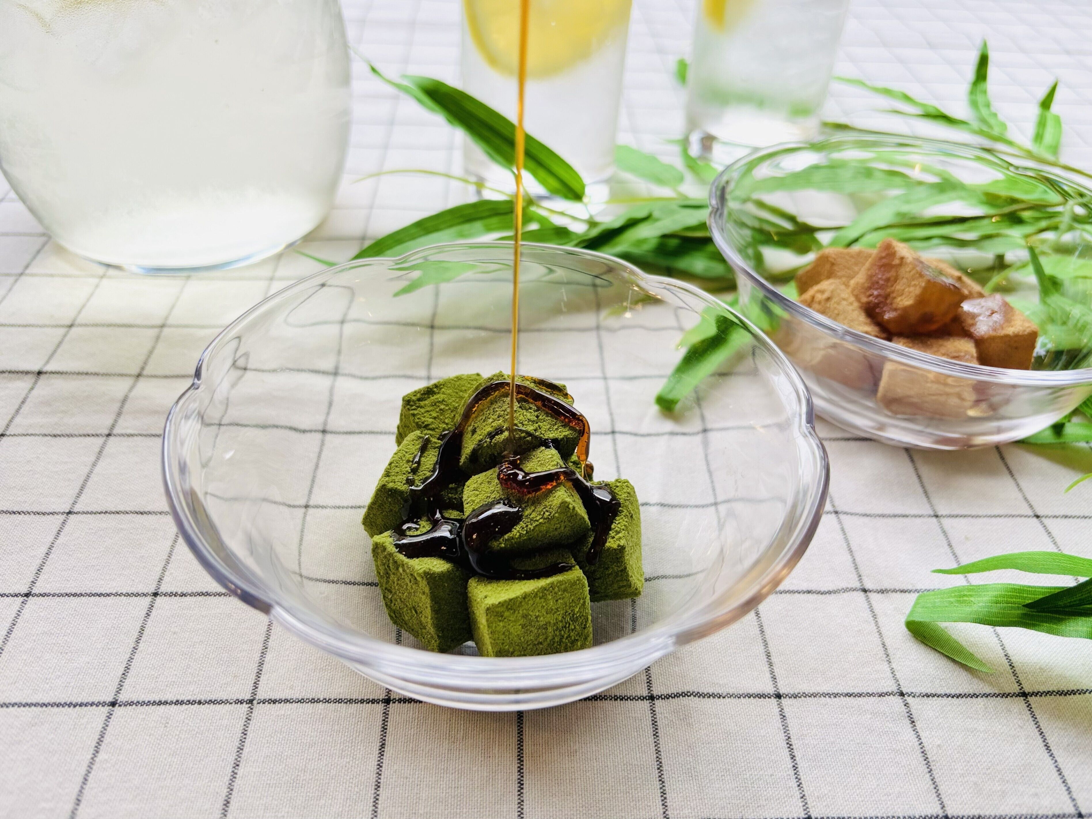 Un tazón de vidrio transparente con cubitos de hielo de matcha y un hilo de jarabe, sobre un mantel a cuadros.