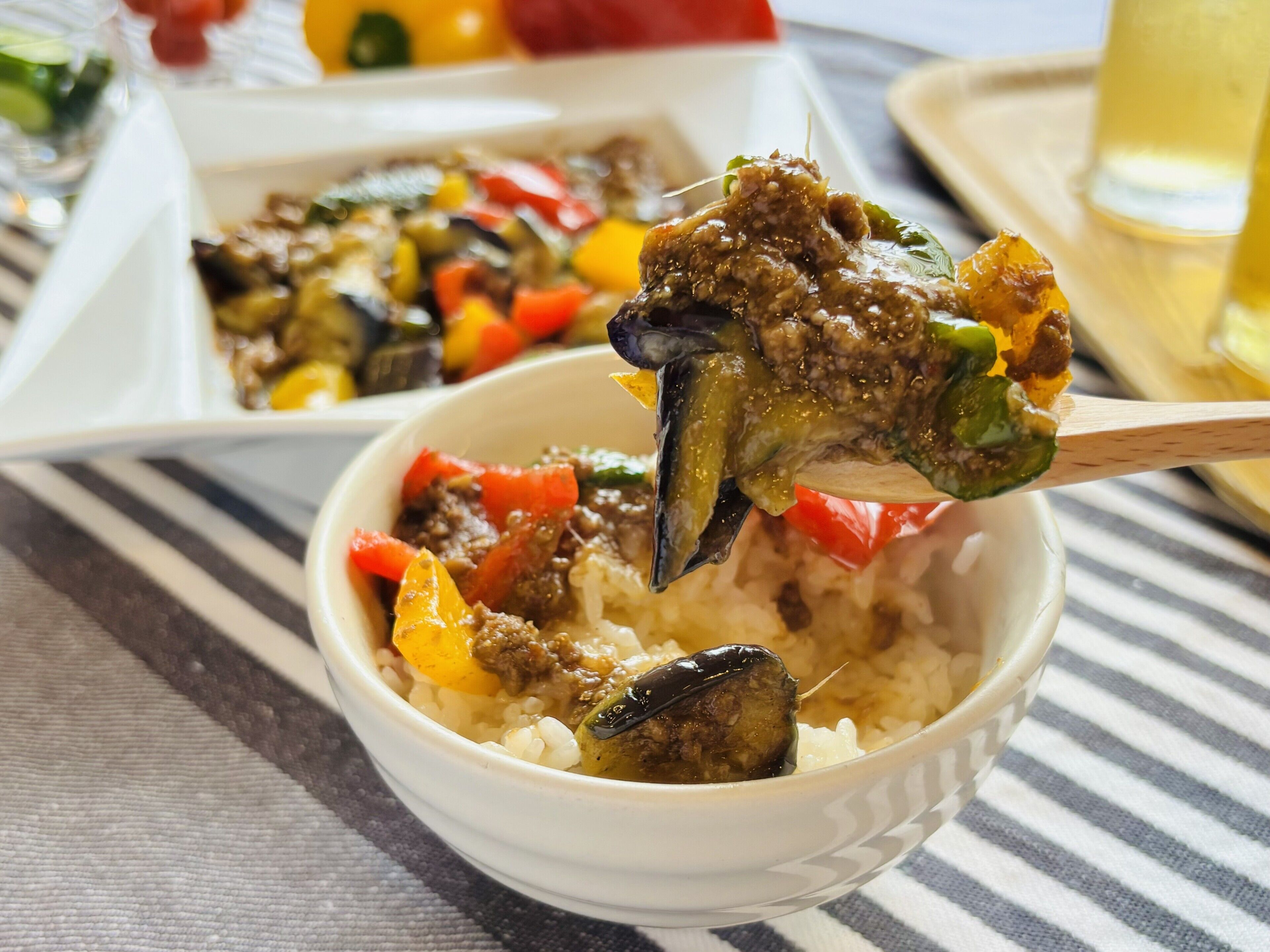 Un tazón de comida con arroz y verduras, una cuchara levantando un bocado, y una bandeja con más comida al fondo.