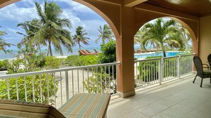 Ein Balkon mit Blick auf den Ozean und Palmen, ein Pool und eine Lounge-Ecke mit gestreifter Sitzkissen.