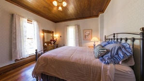 A bedroom with a wooden ceiling, a bed with a white bedspread, a nightstand with a lamp, and a window with curtains.
