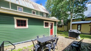 A green house with a red door, a covered grill, and a table with chairs in the yard.