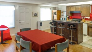 A dining area with a red table and four chairs, a kitchen with wooden cabinets, and a window with blinds.