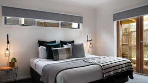 Un dormitorio moderno con una cama grande, varios almohadones, una mesita de noche con lámpara y vistas a una terraza de madera a través de un ventanal.