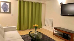 Una sala de estar con un sofá blanco, una mesa de centro de vidrio con un jarrón de flores, una cortina verde, un radiador blanco y un televisor de pantalla plana sobre un soporte de madera.