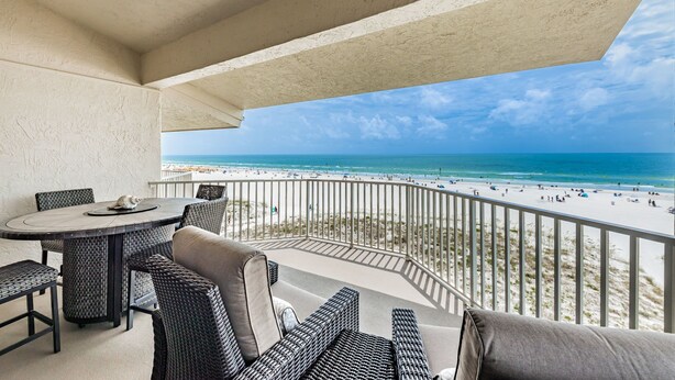 A balcony with wicker furniture, a round table, and a view of the beach and ocean.