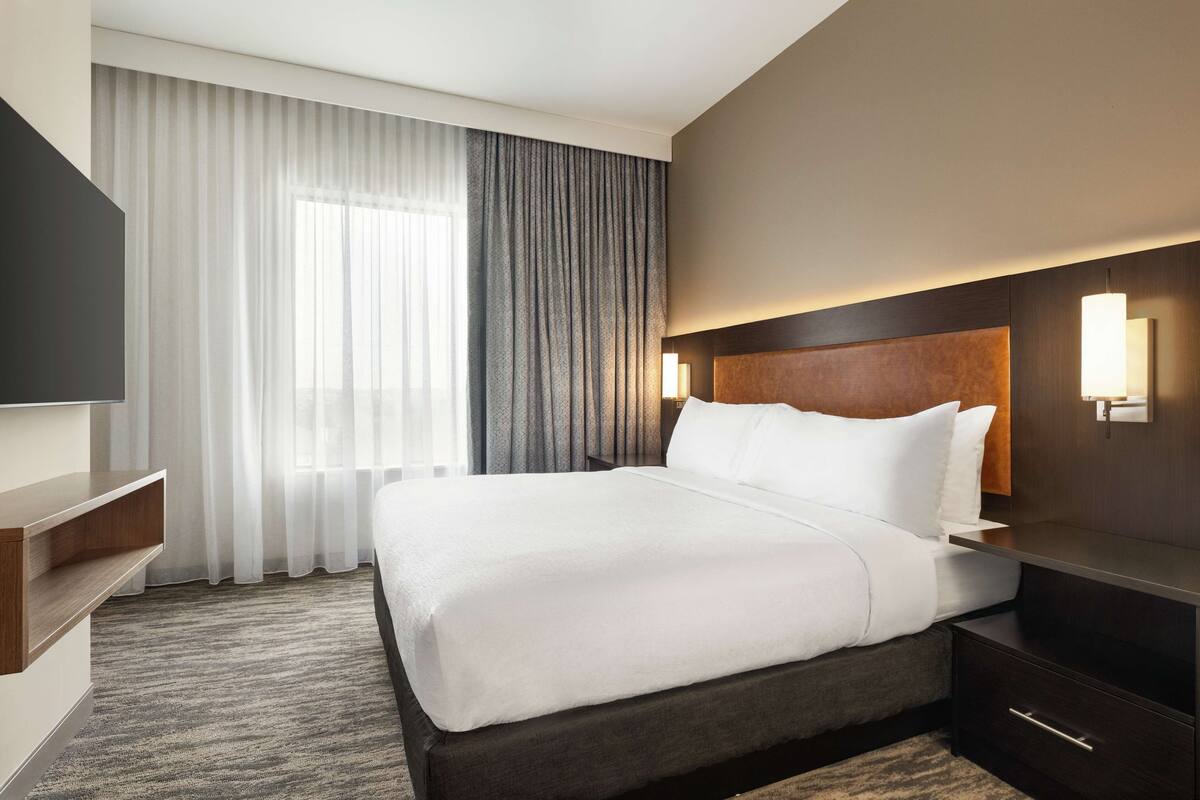 A modern hotel room with a large bed, a flat-screen TV, a bedside table, and a large window with curtains.