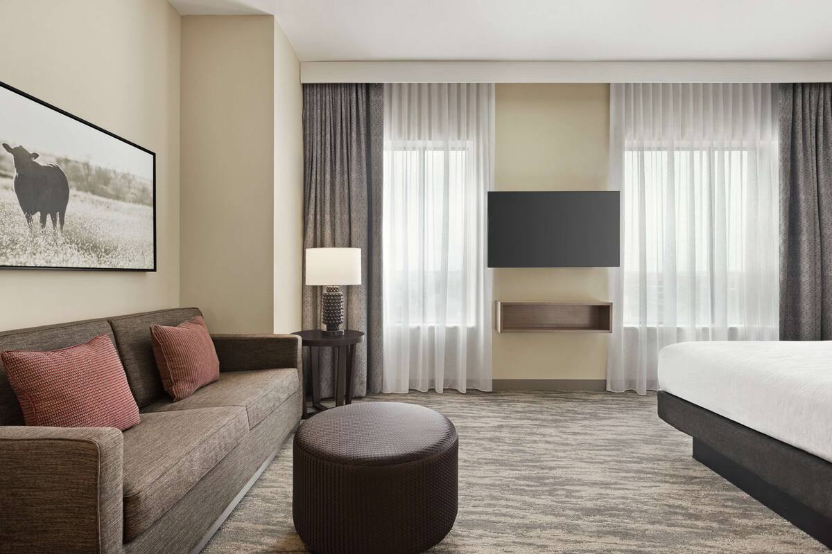 A hotel room with a sofa, ottoman, flat-screen TV, and a large window with curtains.