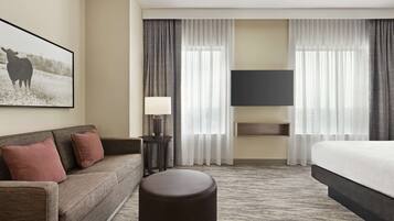 A hotel room with a sofa, ottoman, flat-screen TV, and a large window with curtains.