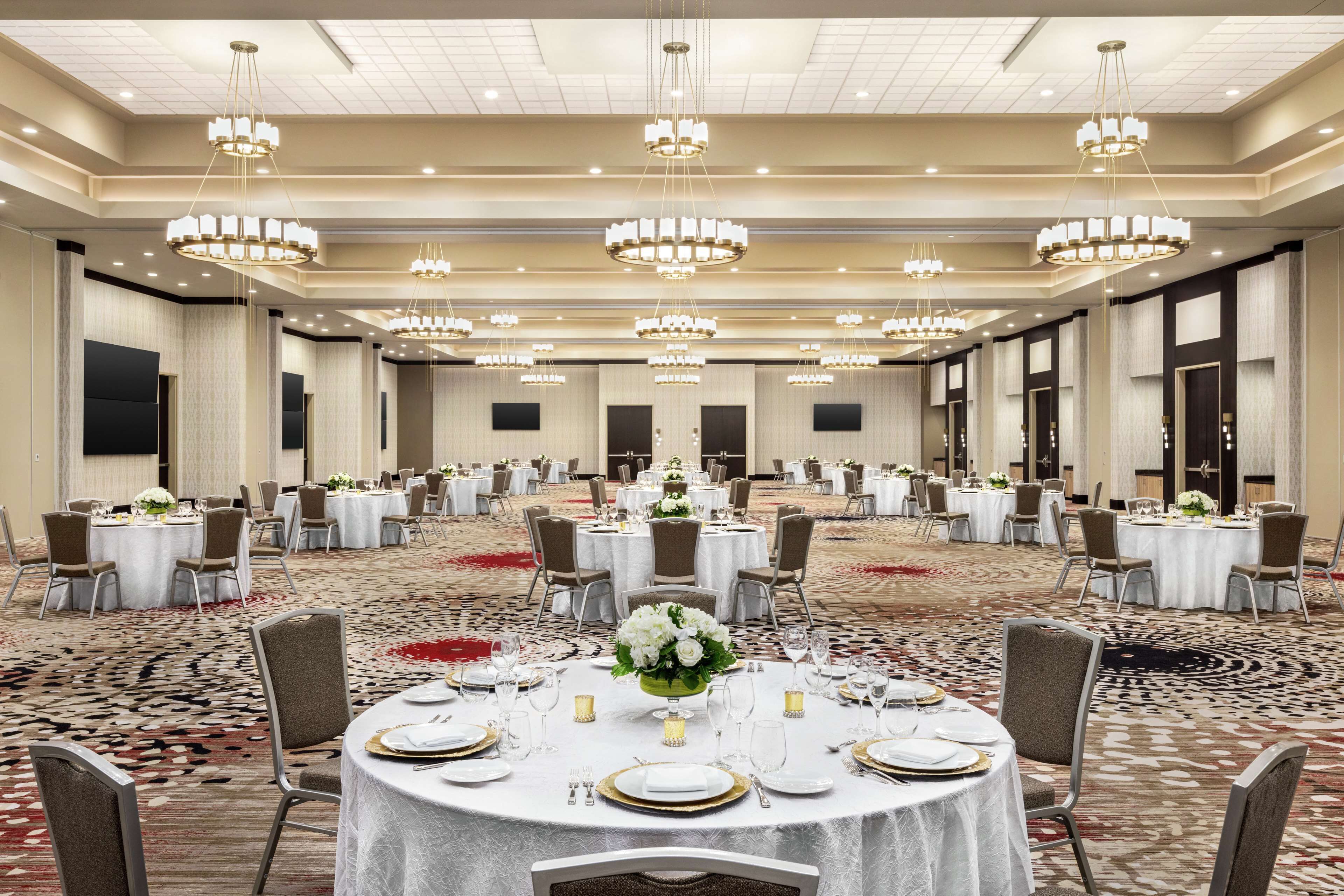 Un amplio salón de banquetes con numerosas mesas redondas dispuestas para comer, cada una con manteles blancos, platos con borde dorado y centros de mesa florales.