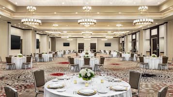 Un amplio salón de banquetes con numerosas mesas redondas dispuestas para comer, cada una con manteles blancos, platos con borde dorado y centros de mesa florales.