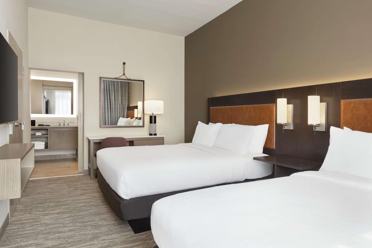 A hotel room with two beds, a desk, a mirror, and a bathroom visible through an open door.