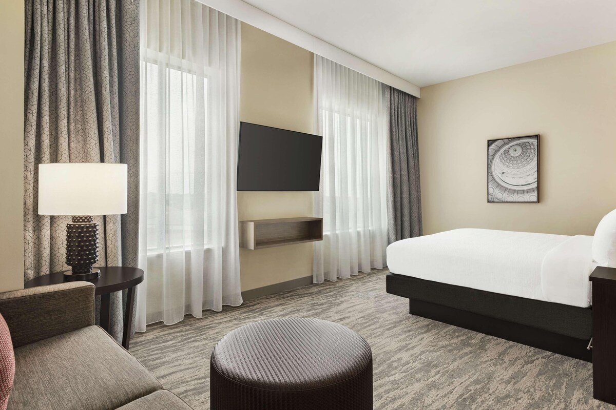 A hotel room with a bed, a TV mounted on the wall, a small table with a lamp, a chair, and a large window with curtains.