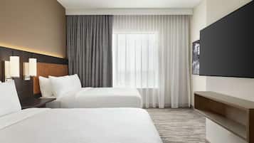 A hotel room with two beds, a large window with sheer curtains, a flat-screen TV, and a wooden shelf.