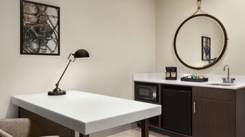 A modern kitchen with a white countertop, a coffee machine, a microwave, and a sink. There is a round mirror on the wall and a framed picture above the countertop.