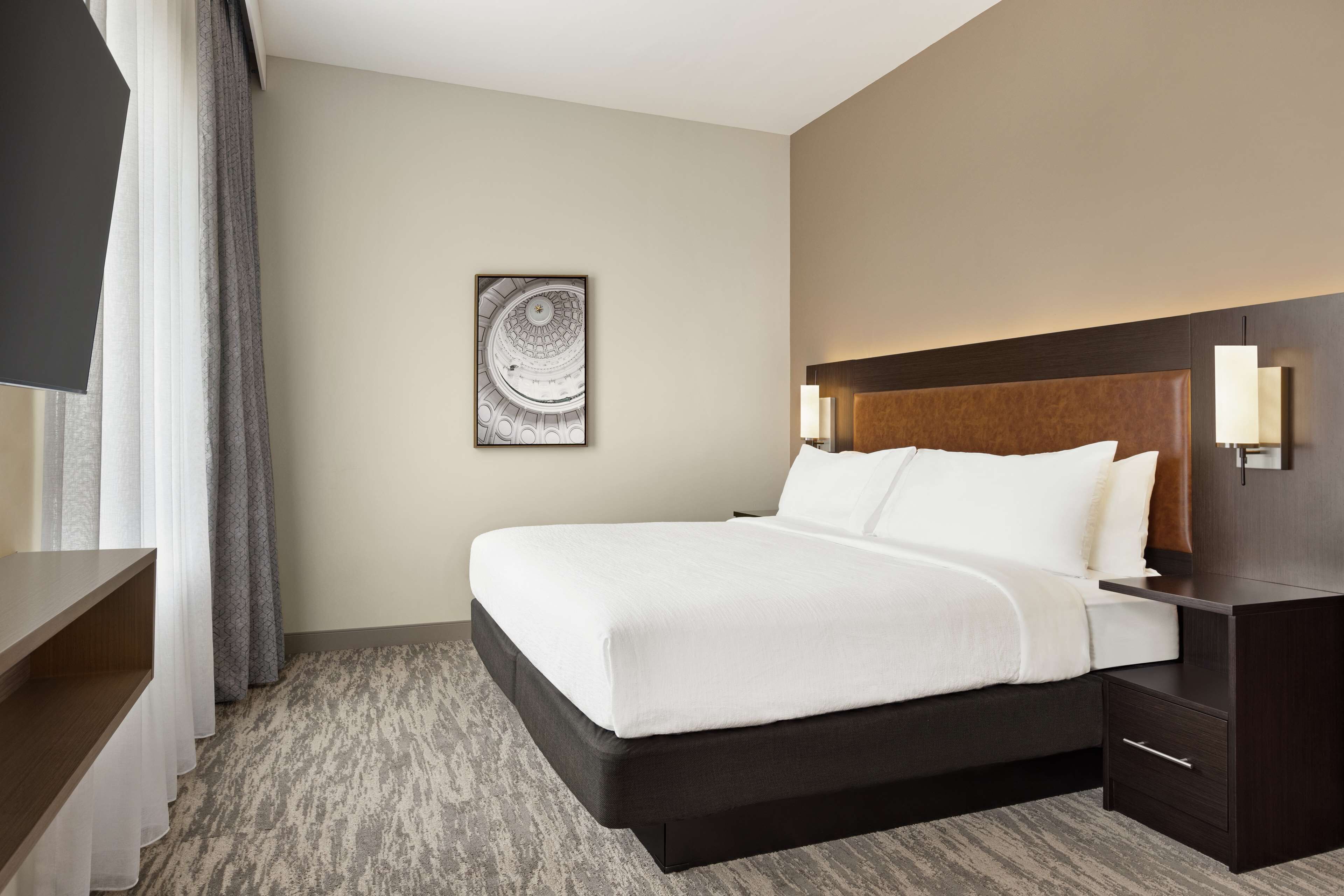 A hotel room with a large bed, a wooden headboard, bedside lamps, a flat-screen TV, and a framed artwork on the wall.