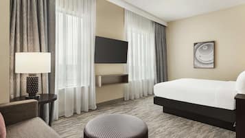 Ein Hotelzimmer mit Bett, Wandfernseher, Nachttisch mit Lampe und großem Fenster mit Vorhängen.