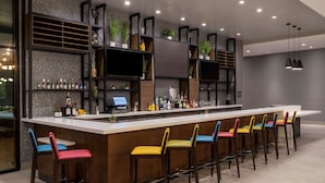 Un espace bar moderne avec un long comptoir, de nombreuses étagères remplies de bouteilles et des tabourets de bar aux couleurs vives.