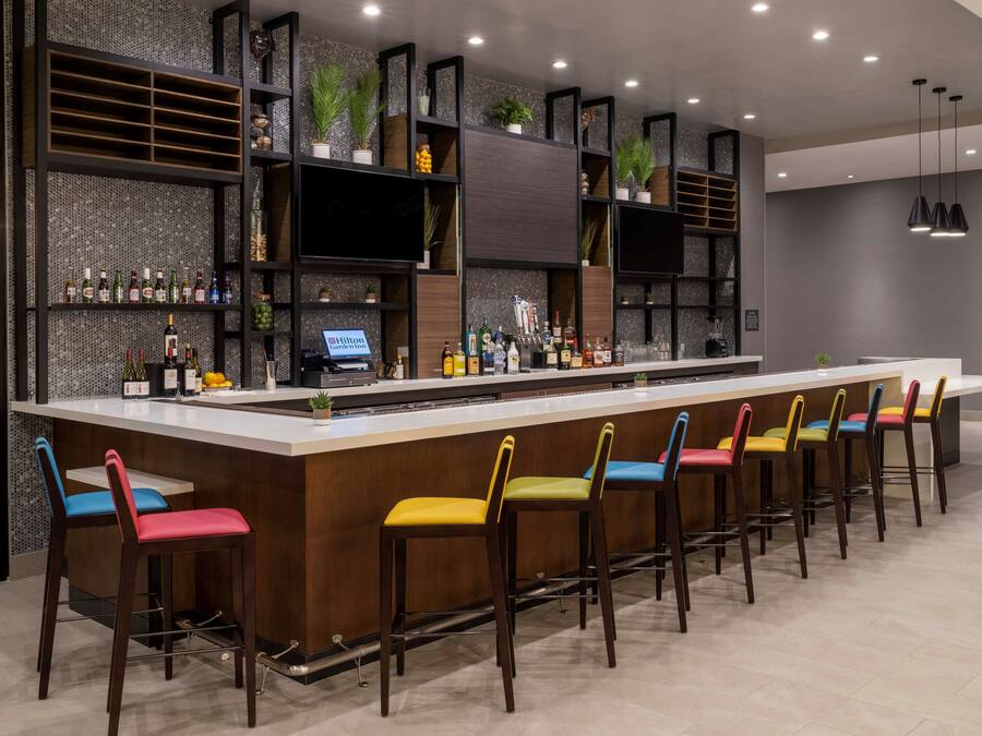 A modern bar area with a long counter, multiple shelves with bottles, and colorful bar stools.